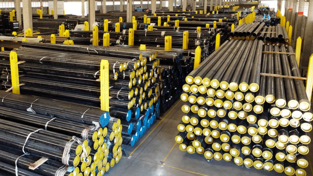 Visão panorâmica de um vasto estoque de tubos de condução de aço carbono, com proteções azuis e amarelas em um grande depósito.