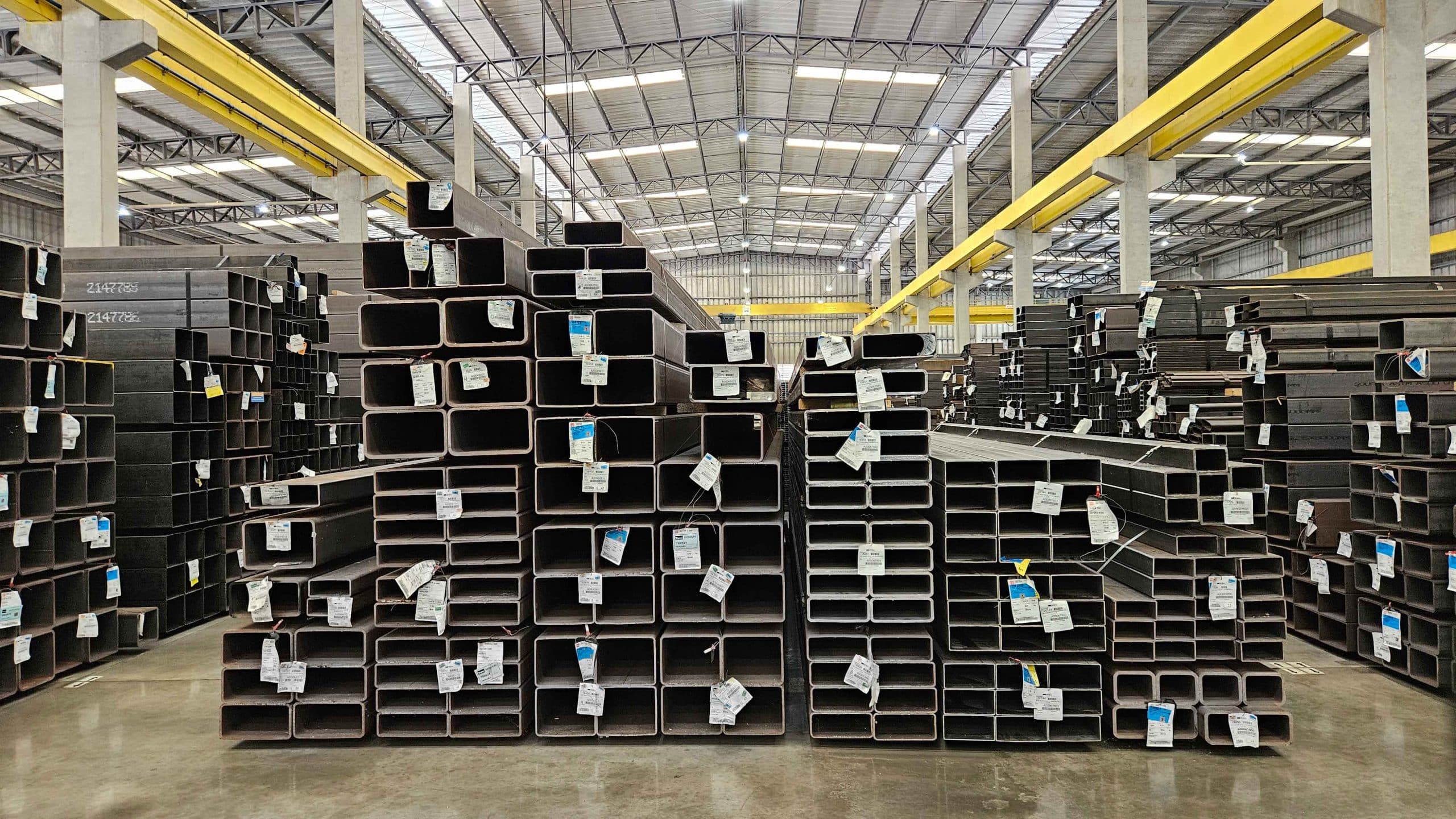 Interior de um grande galpão, exibindo pilhas altas de tubos de aço estruturais retangulares e quadrados, organizados e com etiquetas de identificação.