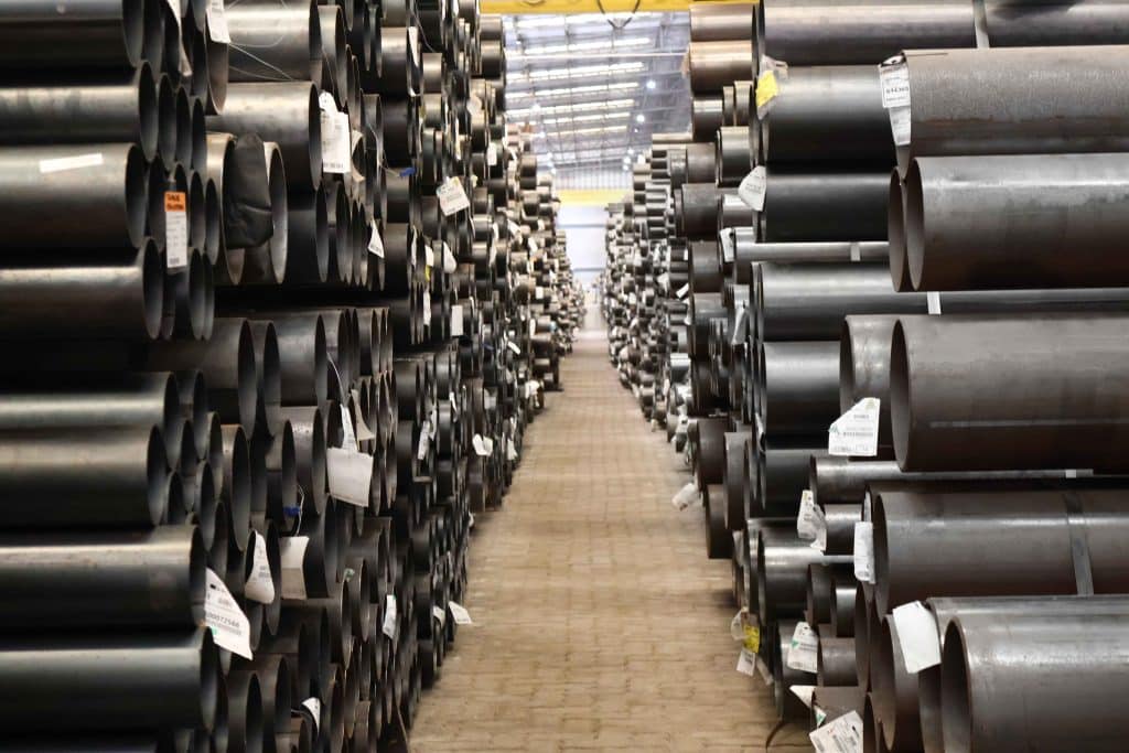 Corredor de depósito industrial com pilhas altas de tubos de aço estruturais redondos de diferentes tamanhos, estocados com etiquetas.