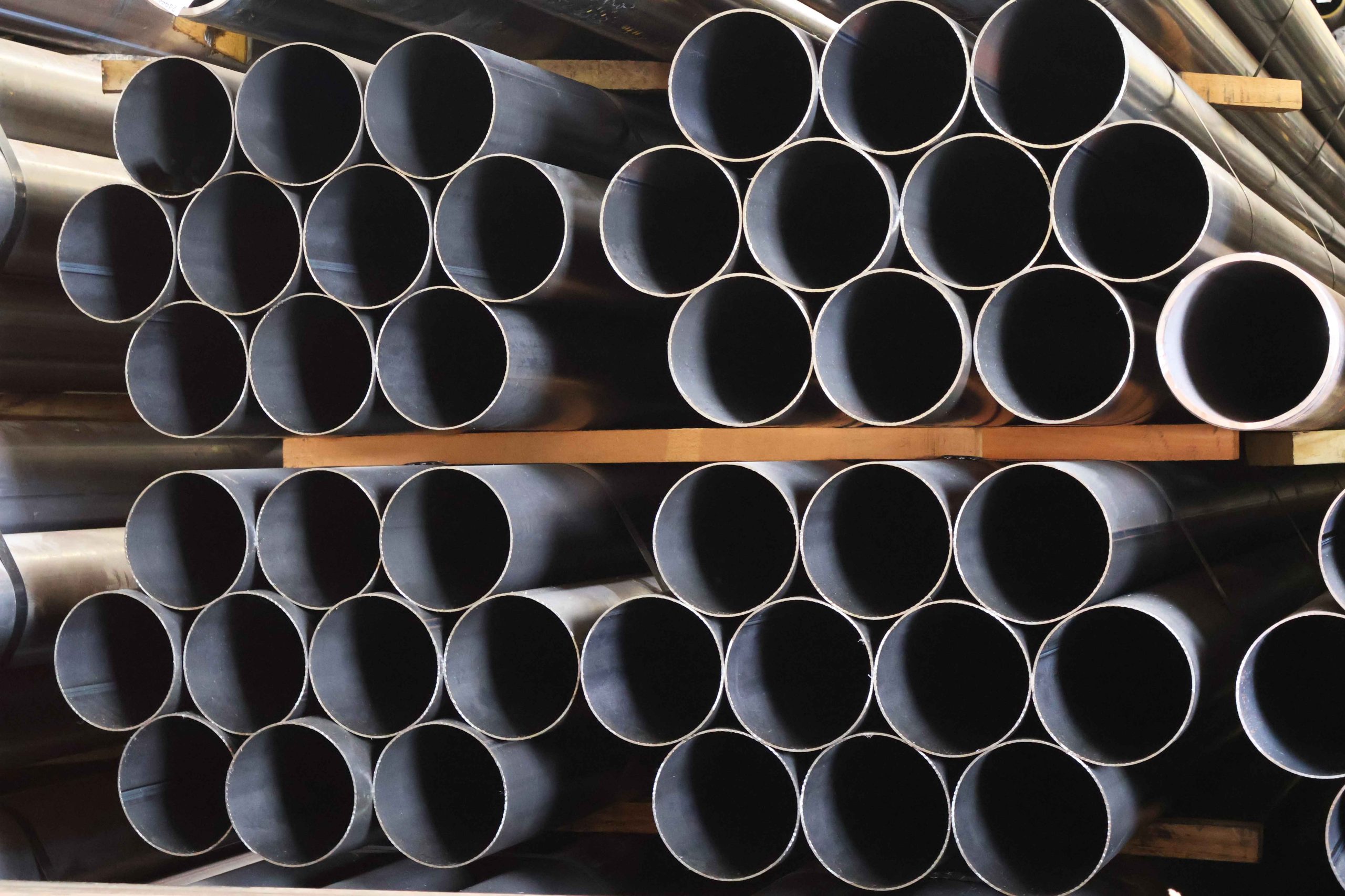 Pilhas organizadas de tubo redondo de aço carbono, separadas por prateleiras de madeira no estoque.
