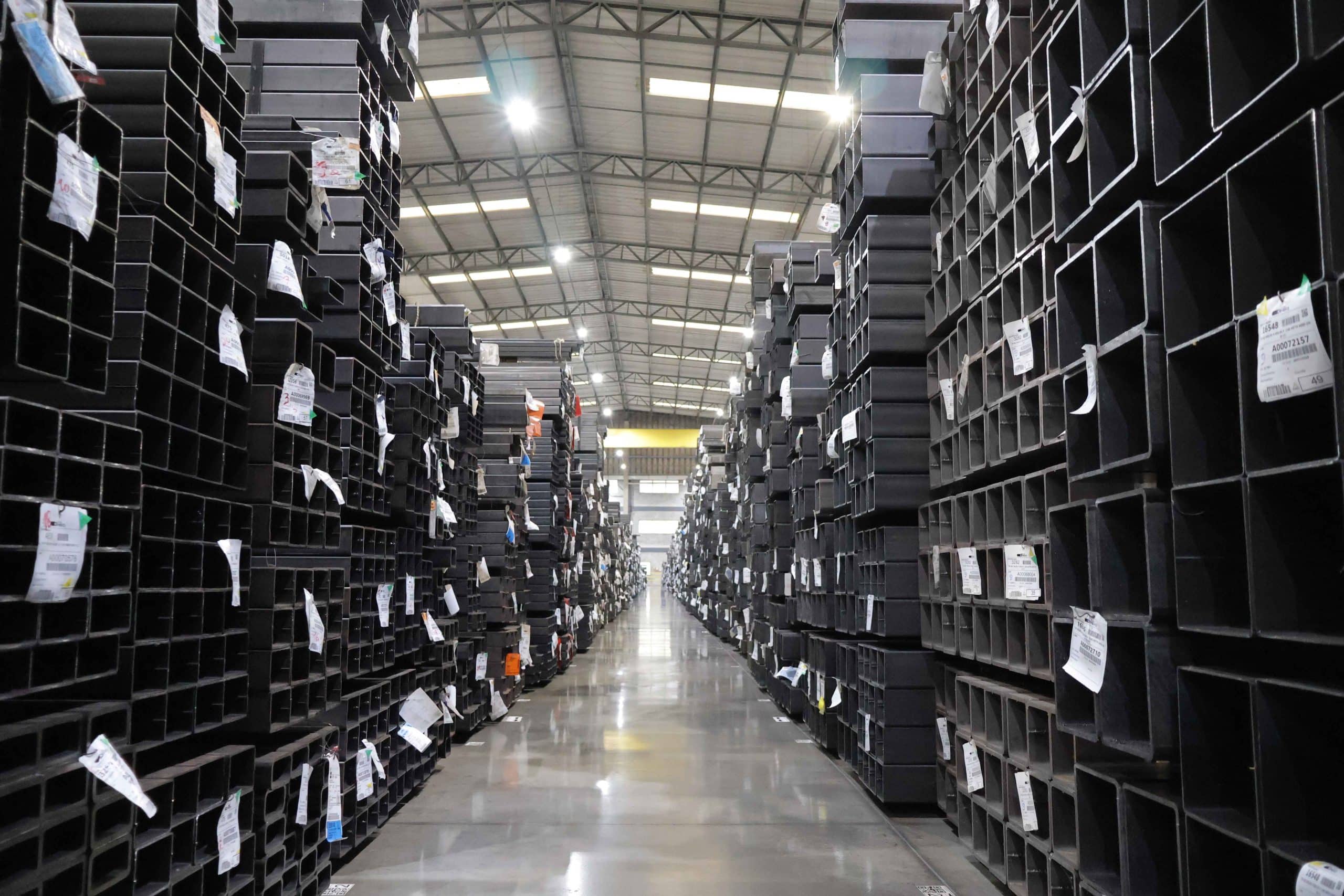 Grandes pilhas de tubos de aço estruturais quadrados de diferentes tamanhos, empilhados e organizados no galpão industrial.