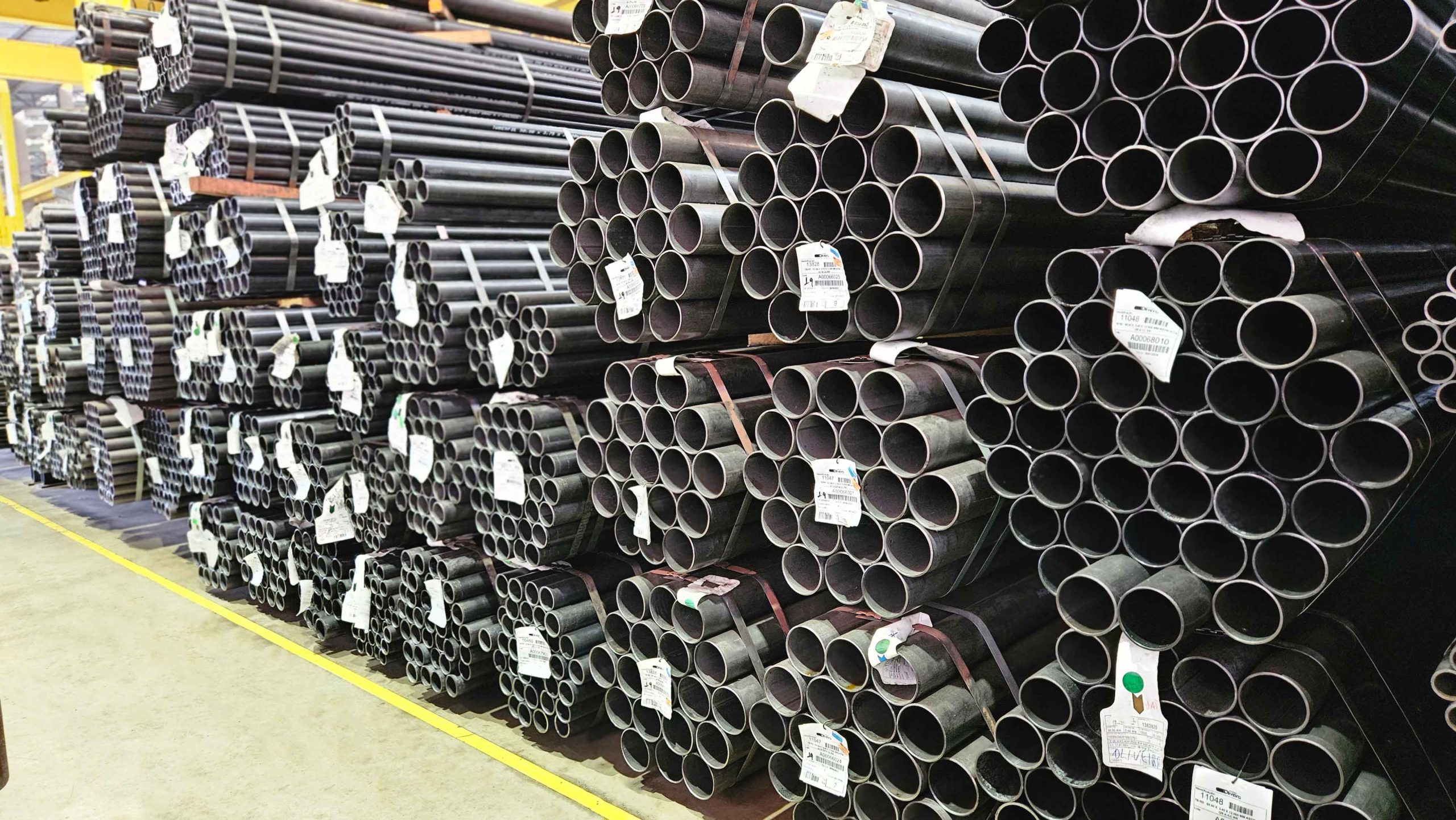 Amplo estoque de tubos redondos de diferentes diâmetros, empilhados e amarrados no galpão industrial.