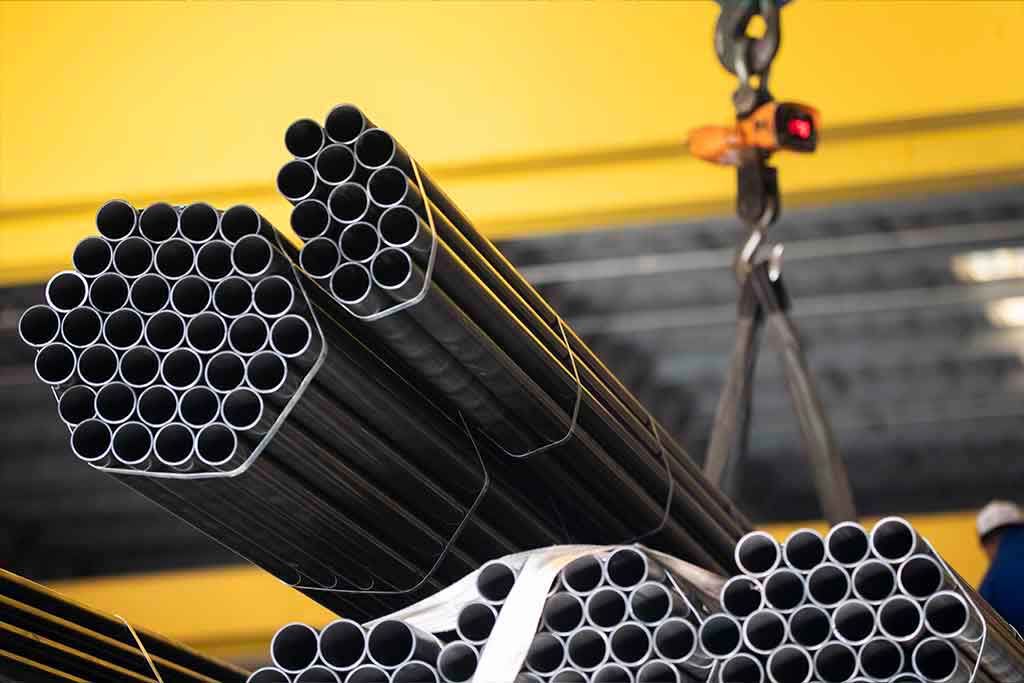 Tubos redondos de aço na balança móvel, içado por gancho em fundo industrial amarelo, pronto para pesagem e expedição.