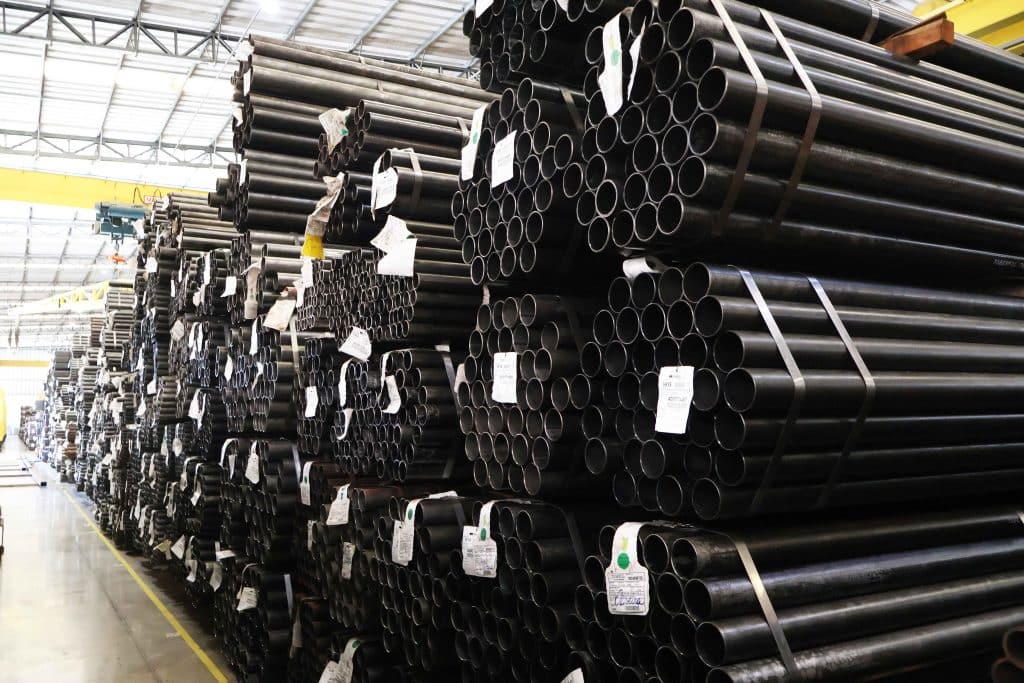 Grandes pilhas de tubos de aço de norma ASTM A178 estocados e amarrados, prontos para distribuição em galpão industrial.