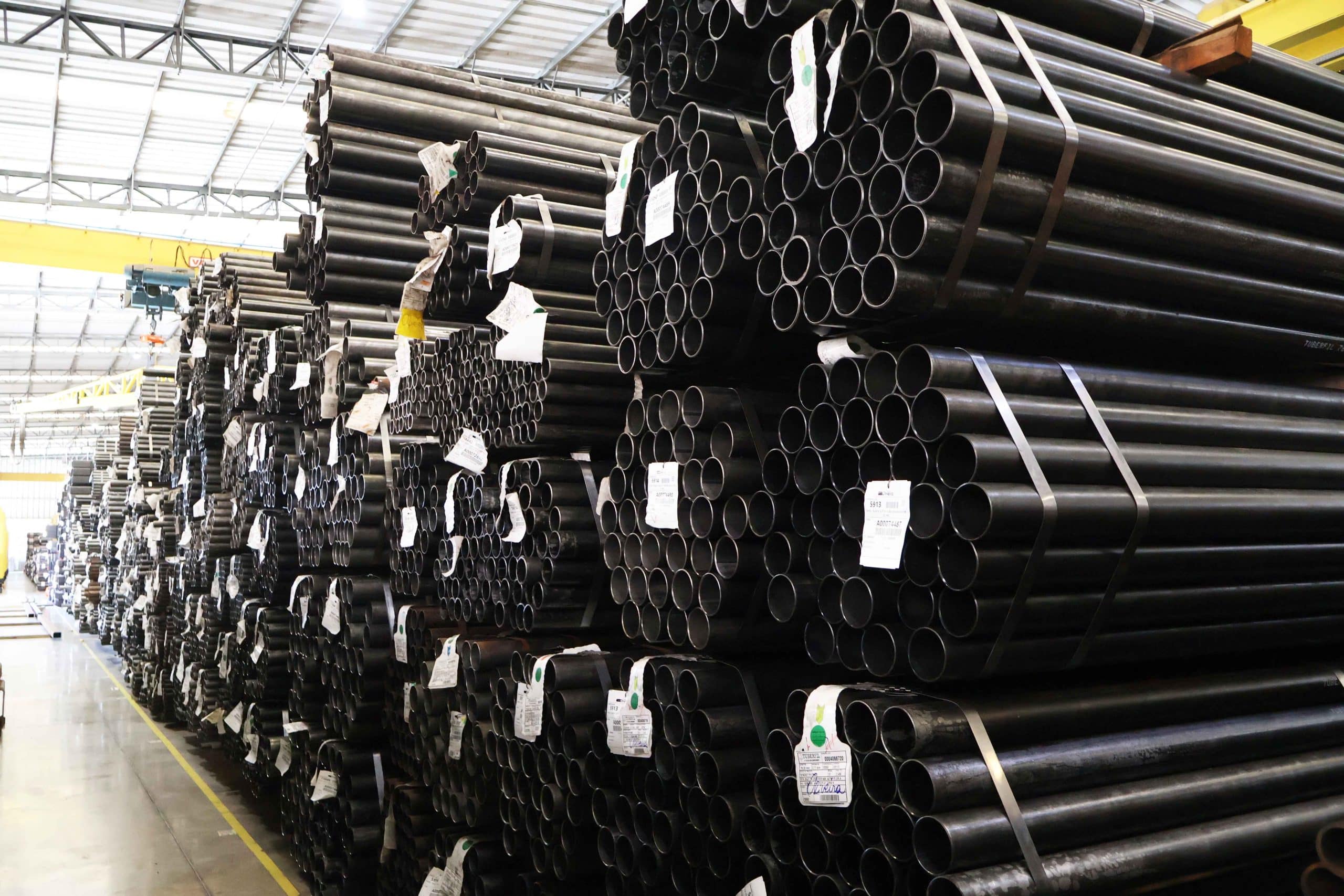 Grandes pilhas de tubos de aço de norma ASTM A178 estocados e amarrados, prontos para distribuição em galpão industrial.