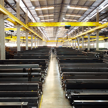 Grande armazém industrial, mostrando um extenso estoque de tubos de aço organizados em pilhas e servido por pontes rolantes.