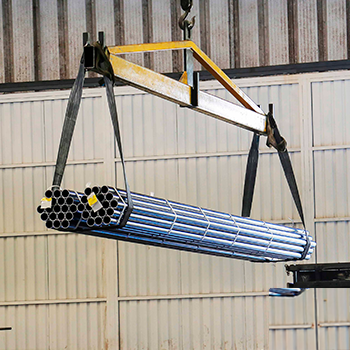 Ponte rolante amarela içando um amarrado de tubos redondos de aço com cintas de elevação, movimentando a carga pesada dentro do galpão industrial.